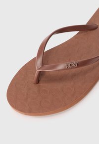 Ruda guminė šlepetė, turinti tekstūruotą vidpadį su apskritais raštais ir plonu, kreiviu dirželiu. Roxy logotipas iškaltas ant dirželio.