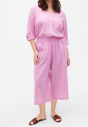 Kvinde iført løs, lyserød skjorte med knapper og opkaldte ærmer samt matchende brede, korte bukser, kombineret med brune åbne slip-on sandaler.