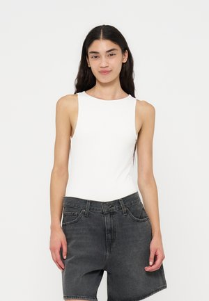 Personne aux longs cheveux foncés portant un haut blanc sans manches et un short en jean noir, debout devant un fond blanc uni, faisant face en avant.