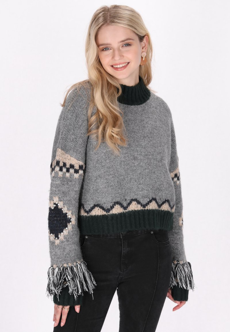 Grauer Wollpullover mit einem gerippten grünen Rollkragen, verziert mit beigen und dunklen Akzenten. Fransen an den Ärmeln verleihen der Textur mehr Tiefe.