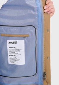 Hnědá voděodolná bunda má vnitřek s modrou podšívkou, kapsu na zip a štítek s textem ve francouzštině a angličtině. Zipper je na boku.