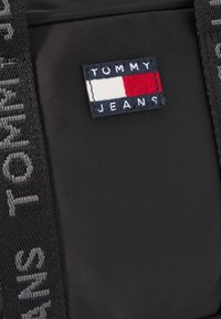 Zwarte stoffen tas met een rechthoekig Tommy Jeans logo patch in marineblauw, rood en wit, en een gestructureerde schouderband met de tekst "TOMMY".