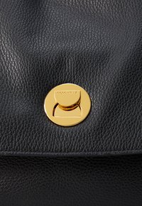 Sac en cuir noir texturé avec un fermoir rond en métal doré présentant le nom de la marque "Coccinelle" gravé. Fini lisse, design épuré.