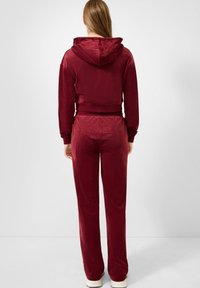 Burgunderfarbene Velour-Jogginganzug mit verkürztem Reißverschluss-Hoodie und passenden Hosen, mit erhabenem Logo auf der Rückseite des Bundes. Glatte Textur.