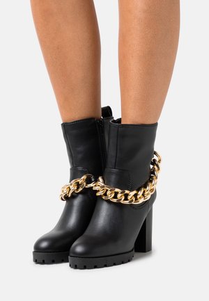 Black leather ankle boots feature a chunky heel and a distinctive gold chain detailing around the ankle. Textured sole for traction.