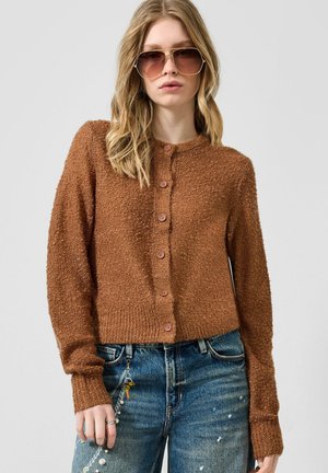 Jeune femme portant un cardigan en maille marron boutonné, un jean bleu avec une chaîne décorative et des lunettes de soleil surdimensionnées, posant devant un fond uni.