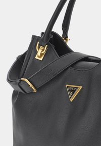 Musta tekstuurinen nahkalaukku, jossa on säädettävä hihna, kultaiset yksityiskohdat ja kolmion muotoinen Guess U.S.A. -logo-merkki edessä.