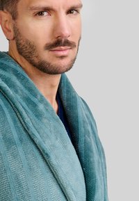 Primer plano de un hombre con cabello corto y oscuro y barba, vestido con un albornoz turquesa contra un fondo gris claro liso.