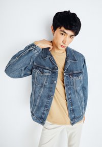 Chaqueta de mezclilla en azul oscuro con diseño clásico, cierre de botones, dos bolsillos en el pecho y una textura ligeramente desgastada. Lleva puesta una camisa beige.
