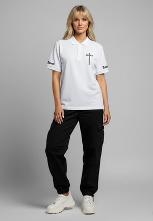 Polo shirt bianca con colletto, caratterizzata da testo grafico nero sulle maniche. Abbinata a pantaloni cargo neri e sneaker bianche e voluminose.