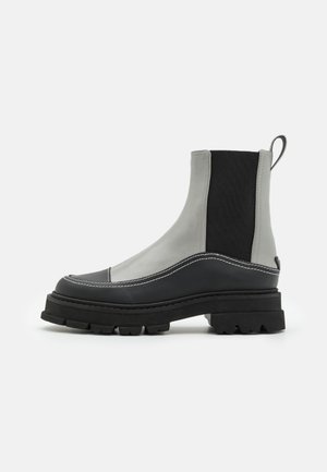 Bottes cheville asymétriques en cuir gris avec un panneau élastique noir. Semelle en caoutchouc épaisse avec un motif de crampons et une finition de coutures blanches.