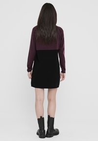 Strickkleid mit maroonem Oberteil und schwarzem Unterteil. Lange Ärmel und glatte Textur. Getragen mit schwarzen Knöchelstiefeln.