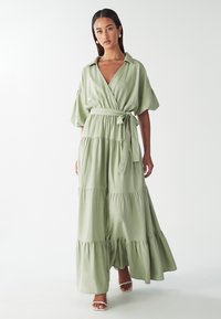 Svetlozelené maxi šaty s preloženým dizajnom, krátkymi naberanými rukávmi, viazaným pásom a tiered volánmi na leme. Hladká, priedušná tkanina.
