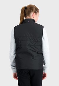 Gilet rembourré noir avec un col haut, présentant des lignes épurées et une texture lisse, porté par-dessus une chemise grise à manches longues.