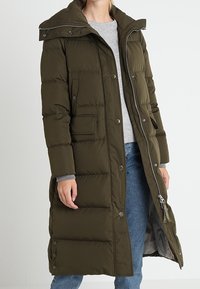 Lang olivengrøn pufferjakke med trykknapper, sidelommer og hætte som kan tages af. Har quiltet struktur og lynlåslukning foran.
