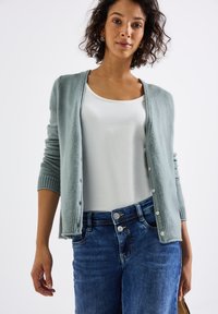 Cardigan vert clair avec une texture douce et des poignets côtelés, fermeture à boutons, associé à un haut blanc et un jean bleu. Design simple et décontracté.