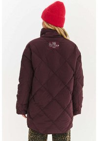 Kurtka puchowa w ciemnym burgundzie z przeszyciami w kształcie diamentu, wysokim kołnierzem i naszywką z logo na plecach, zestawiona z spodniami w panterkę.