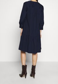 Mörkblå klänning med långa ärmar, volangkjol och rynkad bakdel. Bärs med bruna klackade ankelstövlar. Slät tygtextur.