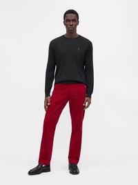 Мъж стои, облечен с черен пуловер с дълги ръкави, червени панталони и черни официални обувки на обикновен бял фон.