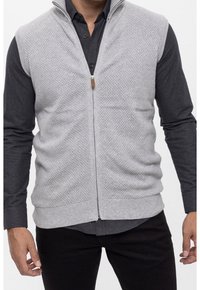 Hellgrauer ärmelloser Pullover mit strukturierter Musterung, Frontreißverschluss und braunem Zipper, getragen über einem dunklen Hemd mit Knöpfen.