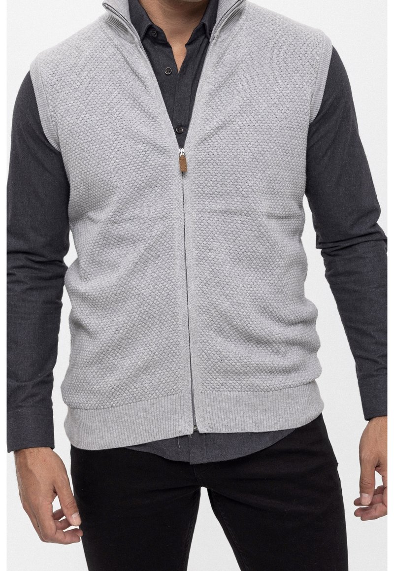 Hellgrauer ärmelloser Pullover mit strukturierter Musterung, Frontreißverschluss und braunem Zipper, getragen über einem dunklen Hemd mit Knöpfen.