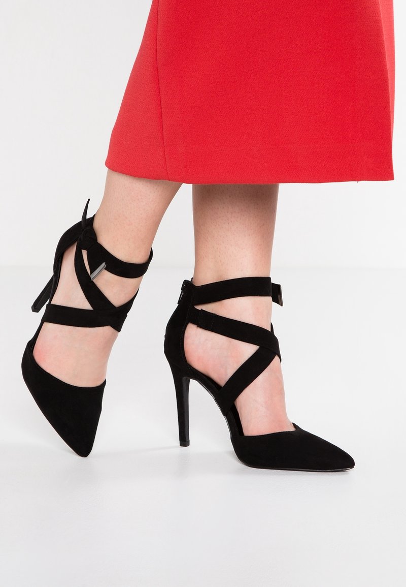 Jambes portant une jupe rouge et des escarpins en daim noir avec des brides croisées à la cheville et des bouts pointus sur un sol blanc.