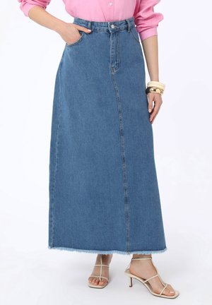 Donna che indossa una lunga gonna di denim blu con orlo sfrangiato, camicia rosa a maniche lunghe, sandali beige con tacco e cinturini, e braccialetti al polso destro.