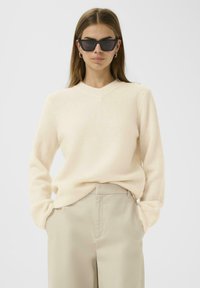Pull beige à col en V en matière douce, accompagné d'un pantalon beige clair. Modèle portant des lunettes de soleil noires angulaires.