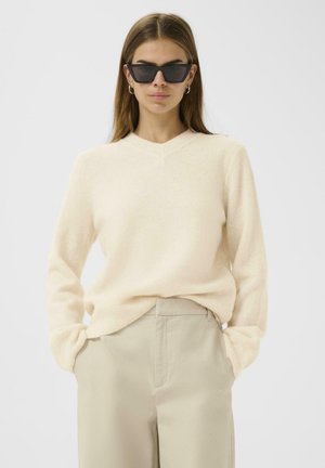 V-neck beige sweater made of soft material, paired with light beige trousers. Model wearing angular black sunglasses.