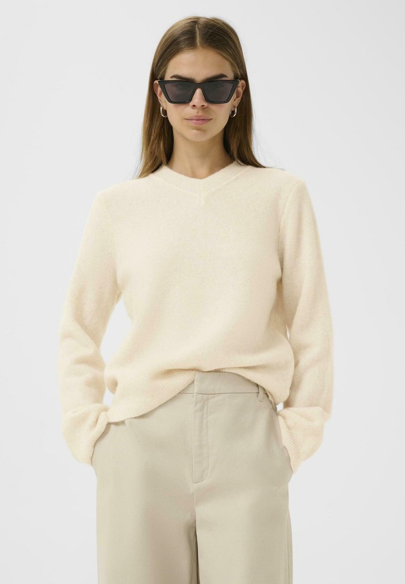 Pull beige à col en V en matière douce, accompagné d'un pantalon beige clair. Modèle portant des lunettes de soleil noires angulaires.
