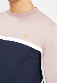 Sudadera de cuello redondo con un diseño de bloques de color en rosa, marino y blanco. Incluye un pequeño logo amarillo en el pecho. Textura de tela suave.