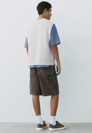 Jonge man staand met zijn rug naar de kijker, draagt een wit gebreid vest, een blauw shirt met korte mouwen, donkere lange korte broek, witte sokken en groene sneakers.