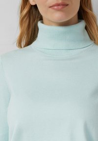 Kobieta w jasnoniebieskim dzianinowym swetrze z golfem, pokazana od szyi do połowy torsu.