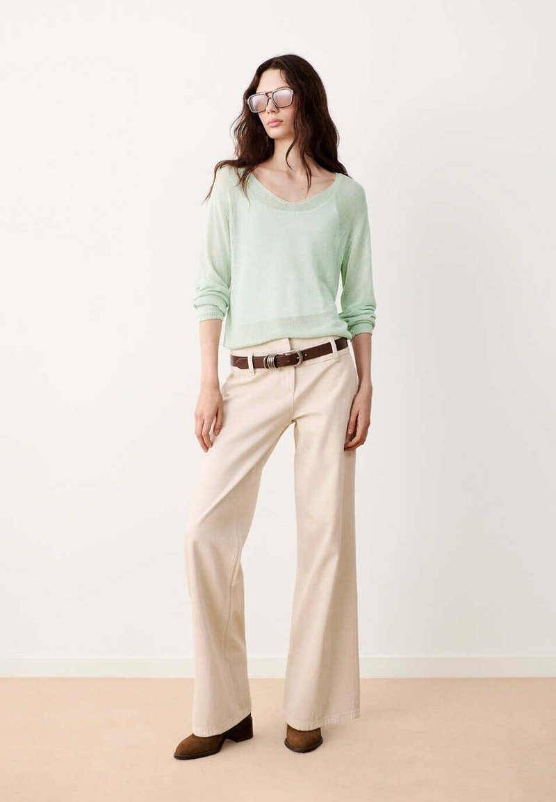 Femme aux longs cheveux foncés portant des lunettes de soleil oversize, un pull vert clair, un pantalon beige à jambes larges, une ceinture marron et des chaussures marron, debout contre un mur uni.