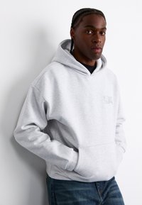 Gray hoodie with a front pocket, cotton material, ribbed cuffs, and a small logo on the chest. Paired with blue jeans, against a white wall.