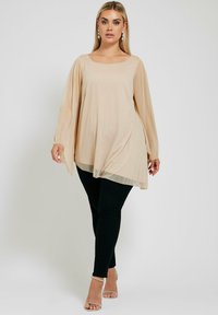 Beige, lätt topp med fyrkantig halsringning, långa ärmar och asymmetrisk nederkant, parat med svarta åtsittande byxor och silverfärgade sandaler.
