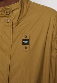 Giacca color khaki con colletto alto, chiusura a bottoni e un'etichetta rettangolare con la scritta "Blauer U.S.A." Il tessuto ha una consistenza liscia.