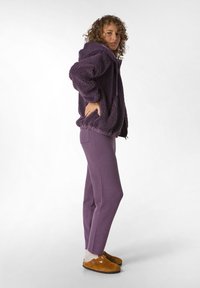 Hanorac din fleece purpuriu cu finisaj texturat, cu glugă ajustabilă și tiv elastic, asortat cu pantaloni fitiți purpurii și saboți maro.