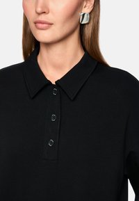 Schwarzes Polohemd mit Kragen, ausgestattet mit einer Dreiknopfleiste. Glatte Textur, aus weichem Stoff gefertigt, kombiniert mit einem glänzenden silbernen Ohrring.