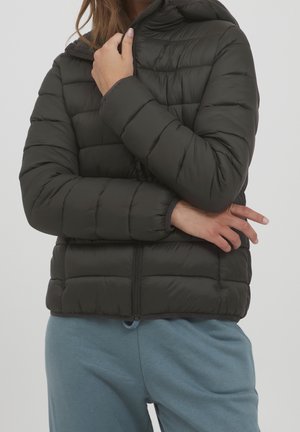 Person iført en sort quiltet dunjakke med hætte, lynet op, og lyseblå bukser, stående med armene krydsede.