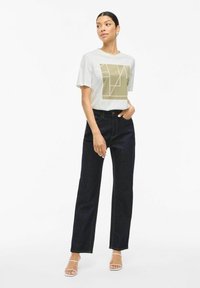 T-shirt i ljusgrått med geometrisk gulddesign, kombinerad med mörkblå raka jeans. Modellen bär genomskinliga klacken sandaler.