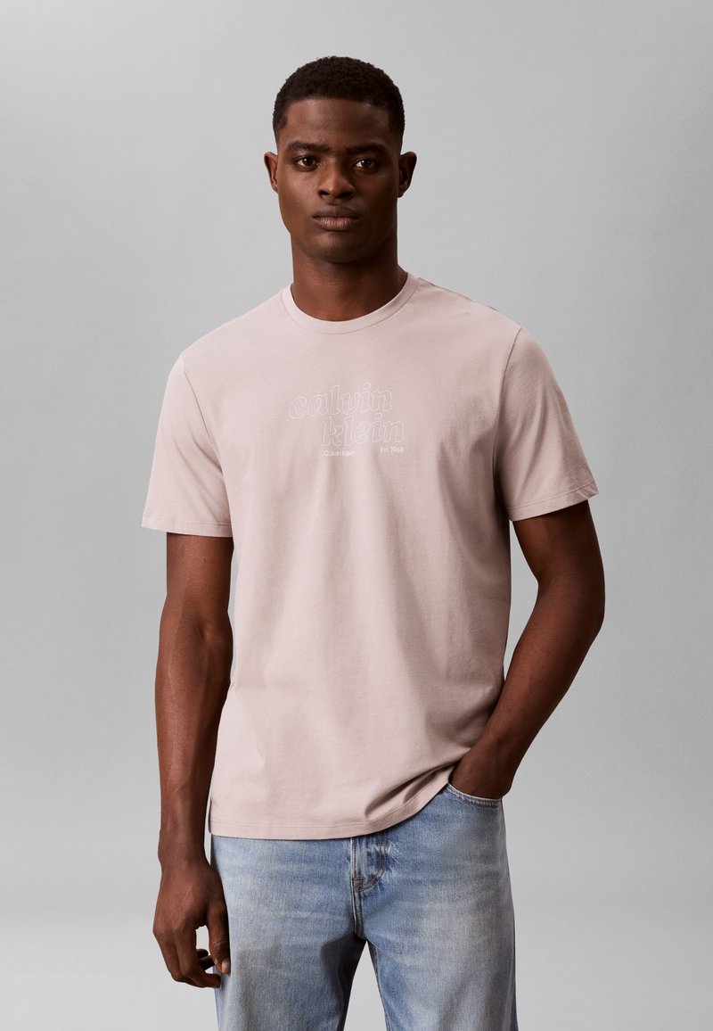 Jeune homme debout avec une main dans la poche, portant un t-shirt Calvin Klein rose clair et un jean bleu clair sur un fond gris uni.