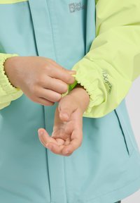 La chaqueta presenta un cuerpo azul claro y mangas de color amarillo neón con puños elásticos. Logo y detalles de texto visibles en la manga. Material texturizado y resistente al agua.