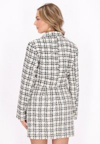 Krém és fekete színű, kockás mintázatú tweed kabát. Hosszú ujjaival, karcsúsított derékrészével és gallér nélküli kialakításával rendelkezik.