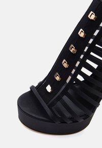 Μαύρη πλατφόρμα μεσαίου τακουνιού με πολλαπλούς ιμάντες και χρυσές τετράγωνες τρουκς κατά μήκος του κάθετου ιμάντα σε λευκό φόντο.