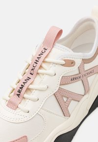 Vit och rosa sportskor med en slät ovandel, texturerade detaljer, synliga snören och en märkt rosa dragflik. Svart yttersula.