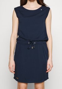 Marinblå klänning med korta ärmar, dragsko i midjan och sidofickor. Har en subtil logotyp vid halsen och en mjuk, jämn textur.