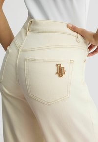 Pantalons taille haute en ivoire avec une finition texturée, présentant un matériel en tone doré sur une poche arrière et des détails de couture visibles.