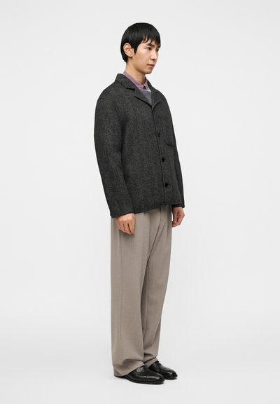 Γκρι υφασμάτινο μάλλινο σακάκι με γιακά, κλείσιμο με κουμπιά και μοναδική τσέπη στο στήθος, φορεμένο πάνω από ανοιχτό μωβ πουκάμισο και μπεζ παντελόνι.