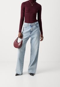 Haut ajusté à manches longues bordeaux, jeans bleu clair à jambes larges, escarpins noirs à talons aiguilles, et un petit sac à main en maroon texturé avec une poignée courbée.